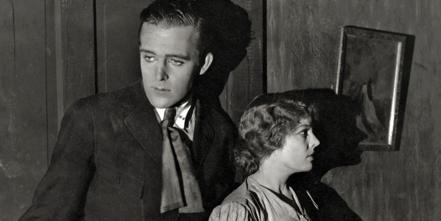 Black-and-white still from "The Cheat" (1915) showing actors Fannie Ward and Jack Dean standing close together in a dim interior, both looking off to the side with tense expressions.
